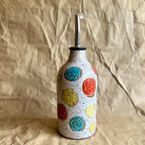 May include: A ceramic oil dispenser with a speckled off-white glaze featuring hand-painted circular motifs in bright blue, yellow, and orange. The bottle has a narrow neck topped with a stainless steel pourer spout for controlled liquid dispensing.