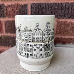 May include: A ceramic cup with a glossy off-white finish featuring a detailed black line drawing of a row of traditional Dutch canal houses wrapping around the exterior. The vessel has a cylindrical upper section and a slightly tapered base.