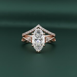 Peut inclure: Une bague de fiançailles en or rose avec une pierre centrale en diamant taille marquise. La bague présente un anneau à tige fendue avec des diamants pavés et un anneau en forme de chevron. La bague est posée sur un fond vert foncé.
