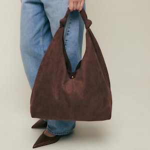 May include: A large, brown suede shoulder bag with a knotted handle. The bag has a simple, unstructured design and a single button closure. The bag is held by a person wearing blue jeans, a white shirt, and a yellow sweater.