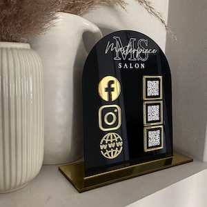 Op de afbeelding: Een boogvormig zwart acrylbord met een gouden voet en de tekst Masterpiece SALON. Het bord toont drie gouden social media iconen aan de linkerkant en drie bijbehorende vierkante QR-codes aan de rechterkant voor Facebook, Instagram en een website.