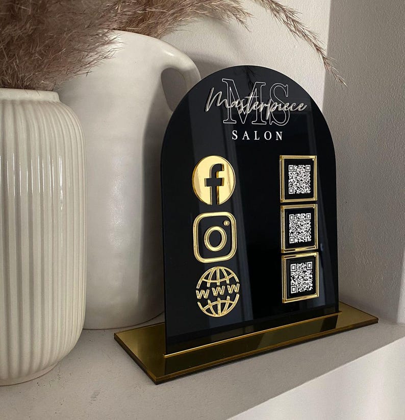 Op de afbeelding: Een boogvormig zwart acrylbord met een gouden voet en de tekst Masterpiece SALON. Het bord toont drie gouden social media iconen aan de linkerkant en drie bijbehorende vierkante QR-codes aan de rechterkant voor Facebook, Instagram en een website.