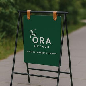 May include: A-frame sign with a dark green panel and black metal frame. The sign features the text "The ORA METHOD" in white, with "PILATES + STRENGTH + CARDIO" below. Brown leather straps secure the panel to the frame.