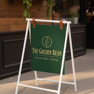 May include: A white metal A-frame sign with a dark green panel. The panel features the text "THE GOLDEN BEAN" in gold lettering, along with a leaf logo. The sign is held in place with brown leather straps.