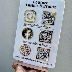 Op de afbeelding: Een wit rechthoekig acrylbord met de tekst Couture Lashes & Browz. Het toont goudkleurige social media iconen voor Instagram, Facebook en een website, elk gekoppeld aan een bijbehorende QR-code en de gebruikersnaam @couturelashesandbrowz.