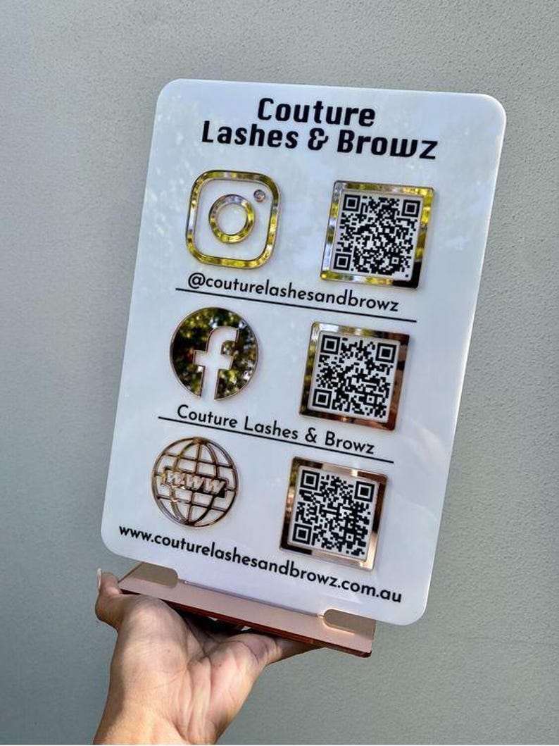 Op de afbeelding: Een wit rechthoekig acrylbord met de tekst Couture Lashes & Browz. Het toont goudkleurige social media iconen voor Instagram, Facebook en een website, elk gekoppeld aan een bijbehorende QR-code en de gebruikersnaam @couturelashesandbrowz.