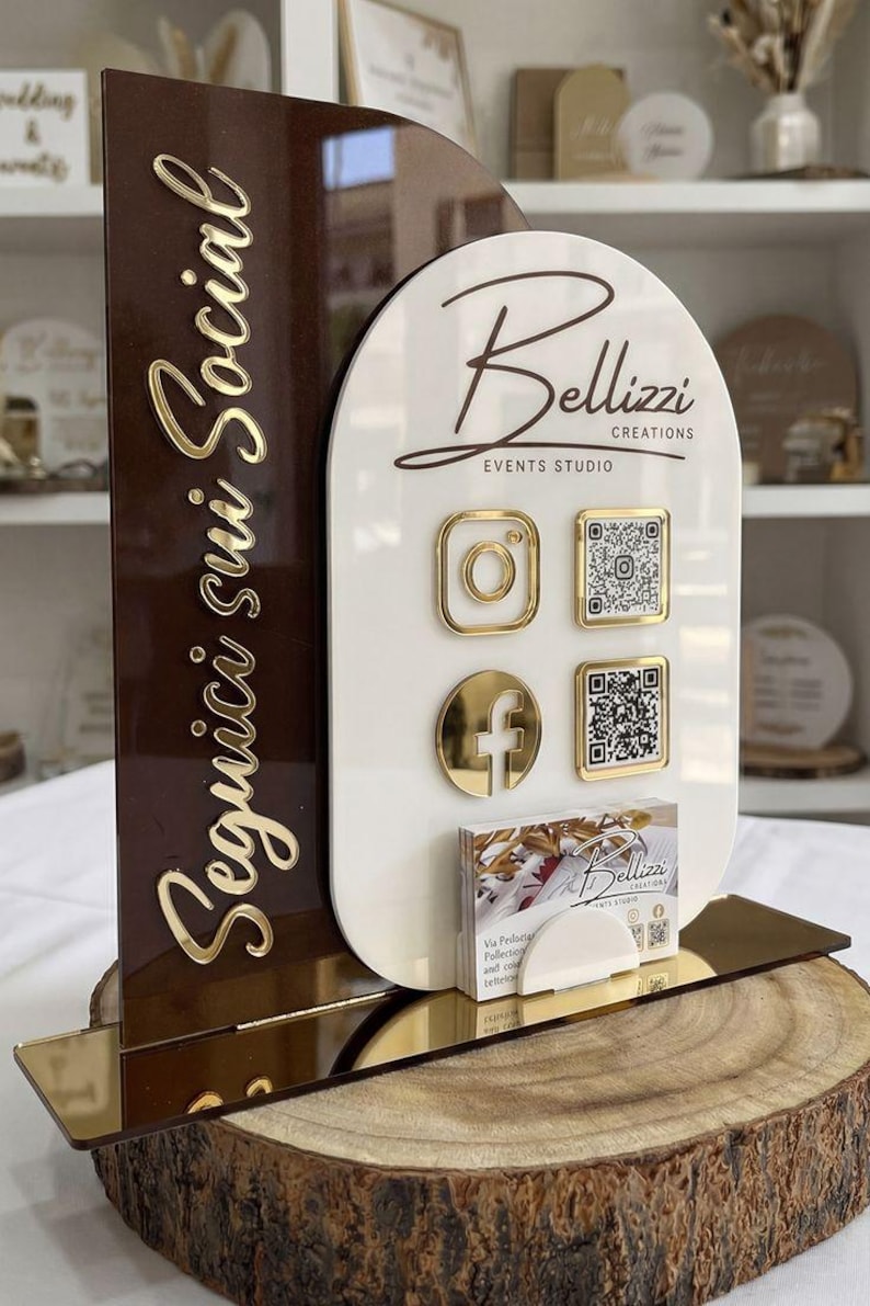 Op de afbeelding: Een dubbelzijdig acryl bord voor sociale media met een donkerbruin boogvormig paneel met gouden tekst Seguici sui Social en een wit paneel met het Bellizzi Creations logo, gouden iconen, QR-codes en een houder voor visitekaartjes.