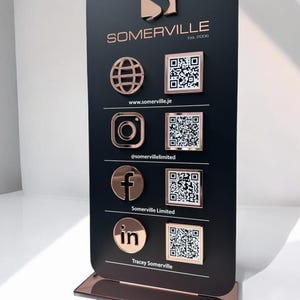 Op de afbeelding: Een verticaal zwart rechthoekig displaybord met afgeronde hoeken, voorzien van ros&eacute;gouden metalen social media iconen en bijbehorende QR-codes. De tekst bevat SOMERVILLE Est. 2006, www.somerville.je, @somervillelimited, Somerville Limited en Tracey Somerville.