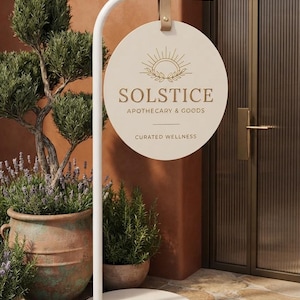 May include: A white metal stand with a circular sign hanging from a tan leather strap. The sign features a sun icon, the text SOLSTICE APOTHECARY & GOODS, and the text CURATED WELLNESS in dark lettering against a cream-colored circular background.