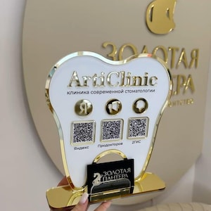 Op de afbeelding: Een tandvormig bord met een witte voorzijde en gouden rand, voorzien van de tekst ArtiClinic, drie ronde iconen en drie QR-codes met de labels Yandex, Prodoctorov en 2GIS. Een klein zwart bordje met gouden tekst is aan de voet bevestigd.
