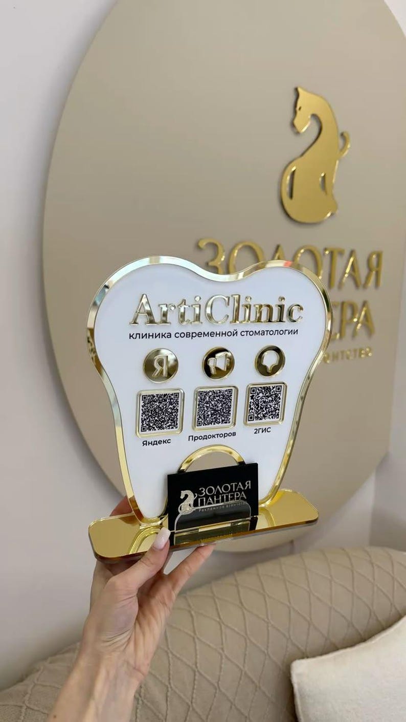 Op de afbeelding: Een tandvormig bord met een witte voorzijde en gouden rand, voorzien van de tekst ArtiClinic, drie ronde iconen en drie QR-codes met de labels Yandex, Prodoctorov en 2GIS. Een klein zwart bordje met gouden tekst is aan de voet bevestigd.