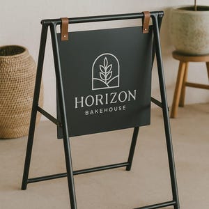 May include: Black metal A-frame sign with the text "HORIZON BAKEHOUSE" in white. The sign has a black background with a white logo of a plant inside an arch. Brown leather straps secure the sign.