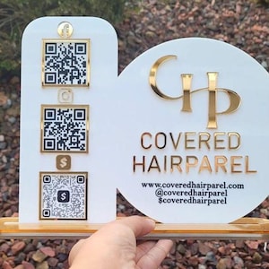 Op de afbeelding: Een wit acrylbord met een verticaal rechthoekig gedeelte met drie goudomrande QR-codes en een cirkelvormig gedeelte met het gouden logo en de tekst COVERED HAIRPAREL, inclusief website en social media gegevens op een gouden voet.