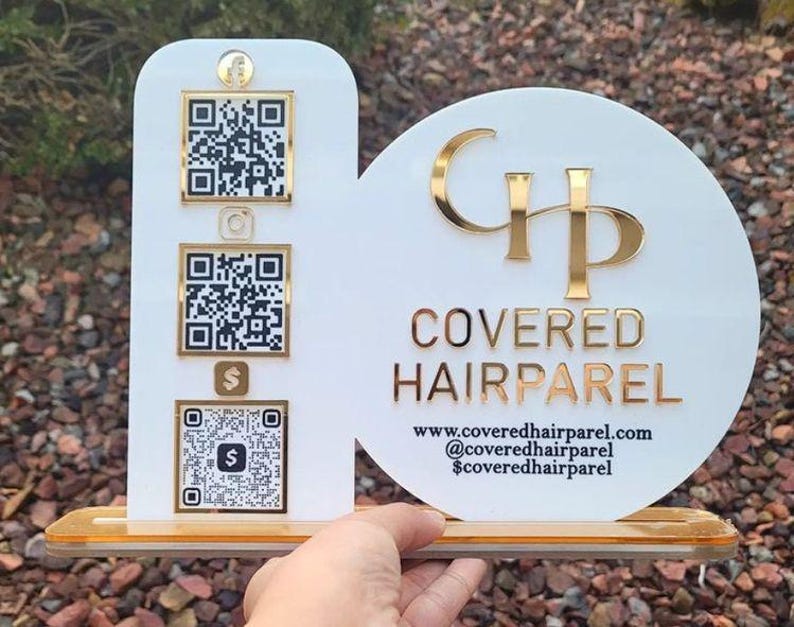Op de afbeelding: Een wit acrylbord met een verticaal rechthoekig gedeelte met drie goudomrande QR-codes en een cirkelvormig gedeelte met het gouden logo en de tekst COVERED HAIRPAREL, inclusief website en social media gegevens op een gouden voet.
