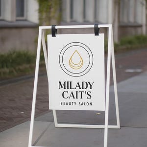 May include: A white A-frame sign with the text "MILADY CAIT'S BEAUTY SALON". The sign features a circular logo with a gold teardrop design. The sign is set up on a sidewalk in front of a building.