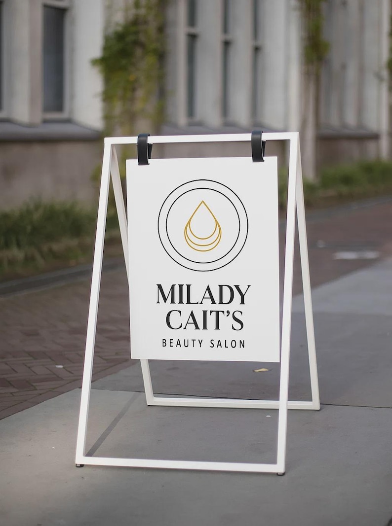 May include: A white A-frame sign with the text "MILADY CAIT'S BEAUTY SALON". The sign features a circular logo with a gold teardrop design. The sign is set up on a sidewalk in front of a building.