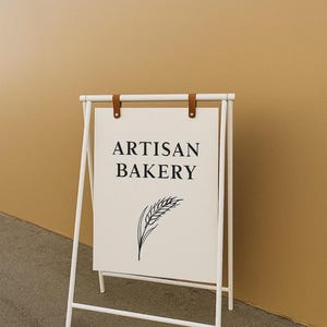 May include: A white A-frame sign with the words "ARTISAN BAKERY" in black, and a wheat stalk illustration. The sign is held by a white metal frame with brown leather straps at the top. The background is a solid tan color.