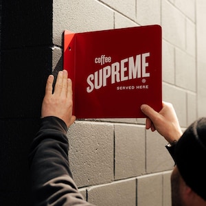 May include: A rectangular red metal sign with a white logo that reads coffee SUPREME SERVED HERE. The sign features a perpendicular mounting flange on the left side, designed to be attached to a wall surface for high visibility signage.