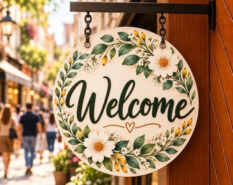 May include: A round, cream-colored hanging sign featuring the word Welcome in dark green script. The sign is decorated with a painted wreath of green leaves, small white flowers, and yellow buds, accented by a small heart symbol below the text.