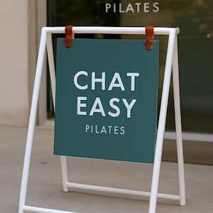May include: A white metal A-frame sign with a teal-colored sign that reads "CHAT EASY PILATES" in white capital letters. The sign is held in place by brown leather straps. The sign is outdoors.