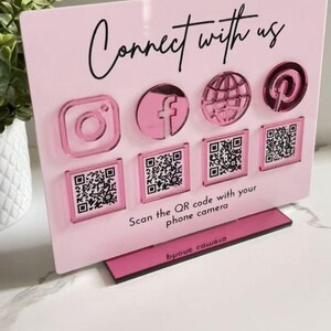 Op de afbeelding: Een rechthoekig lichtroze acrylbord met de zwarte tekst Connect with us en Scan the QR code with your phone camera. Het toont vier verhoogde metallic roze social media iconen en vier bijbehorende vierkante QR-code stickers.