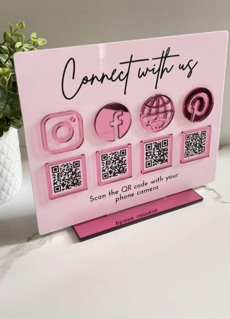 Op de afbeelding: Een rechthoekig lichtroze acrylbord met de zwarte tekst Connect with us en Scan the QR code with your phone camera. Het toont vier verhoogde metallic roze social media iconen en vier bijbehorende vierkante QR-code stickers.