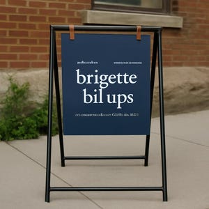 May include: A-frame sign with a navy blue sign featuring the white text "brigette bil ups". The sign is held by a black metal frame with brown leather straps. The sign is outdoors, in front of a brick building.