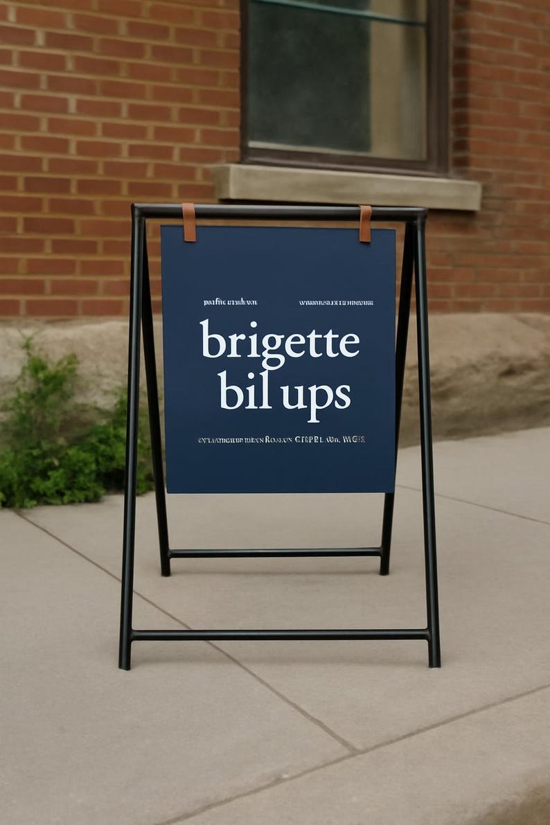 May include: A-frame sign with a navy blue sign featuring the white text "brigette bil ups". The sign is held by a black metal frame with brown leather straps. The sign is outdoors, in front of a brick building.