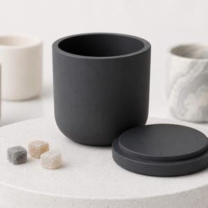 Peut inclure: Un récipient cylindrique noir mat avec un couvercle circulaire assorti, placé à côté de trois petits cubes texturés de forme carrée dans des tons gris et beige, entouré de plusieurs autres récipients similaires aux couleurs neutres.
