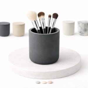 Op de afbeelding: Een donkergrijze cilindrische houder met make-up borstels, geplaatst op een wit rond platform. Verschillende kleine, bijpassende houders in diverse neutrale kleuren zijn op de achtergrond geplaatst. Drie kleine, ronde, pastelkleurige objecten bevinden zich op de voorgrond.