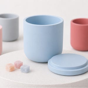 Peut inclure: Un récipient cylindrique bleu clair avec un couvercle circulaire assorti, accompagné de trois petits cubes de sucre translucides et multicolores. L'ensemble est entouré de plusieurs autres récipients cylindriques unis dans des tons rose, gris et vert.
