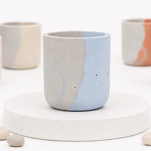 Può includere: Un vaso cilindrico in cemento caratterizzato da un design bicolore con una sezione grigio naturale e una sezione azzurro chiaro. La superficie ha una consistenza opaca con piccole bolle d'aria visibili. Sono disposti diversi vasi simili in tonalità pastello.