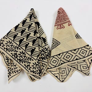 May include: Two folded square cotton napkins in off-white with block-printed designs. One features black geometric zigzags and floral borders. The other displays a red turtle motif at the top with black diamond and triangular patterns along the edges.