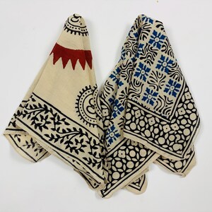 May include: A folded textile featuring a cream-colored base with block-printed patterns. The design includes a red sun-like motif, black floral vines, and a grid of blue floral squares bordered by a repeating black circular and petal pattern.