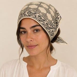 May include: A square headscarf featuring a cream-coloured base with a repeating black geometric pattern and a decorative black floral border. The fabric is tied at the back, displaying a clear contrast between the dark ink and the light textile.