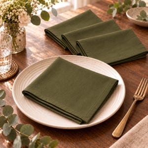 Op de afbeelding: Een set van vier vierkante olijfgroene stoffen servetten, elk met een eenvoudige zoom. Eén servet is netjes gevouwen op een rond, lichtgekleurd keramisch bord, terwijl drie andere bijpassende servetten ernaast op tafel liggen.