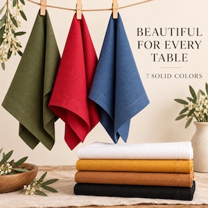 Op de afbeelding: Drie stoffen servetten in olijfgroen, rood en blauw hangen aan een waslijn met houten knijpers. Daaronder liggen vier gevouwen servetten in wit, mosterdgeel, bruin en zwart gestapeld. Tekst: BEAUTIFUL FOR EVERY TABLE 7 SOLID COLORS.