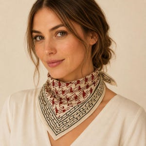 May include: A square neck scarf featuring a cream-coloured base with a repeating pattern of small, dark red abstract shapes. The scarf has a distinct black and cream geometric border design along the edges and is tied in a simple knot at the neck.