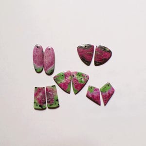 May include: A collection of ten polished ruby zoisite gemstone pendants arranged in five matching pairs. Each piece features a distinct blend of vibrant magenta-pink ruby and earthy green zoisite with black mineral inclusions, all drilled with a small hole.