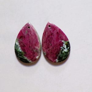 May include: Two teardrop-shaped gemstones with a hole at the top. The stones are a mix of deep pink and green, with a marbled appearance. The gemstones are likely for jewelry making.