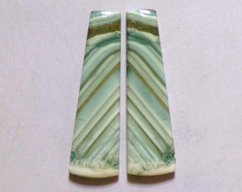 May include: Two elongated, trapezoidal cabochons featuring a pale mint green base color with diagonal, darker green and golden-brown banding. The stones have a smooth, polished surface with a distinct, creamy white textured edge at the base.