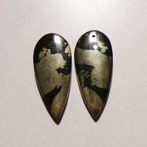 May include: Two teardrop-shaped pendants made of polished pyrite and black stone. Each piece features a smooth, reflective surface with a metallic gold-coloured mineral pattern embedded within the dark stone, and a small hole drilled at the top.