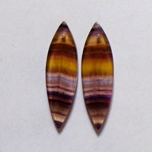 May include: Two elongated, teardrop-shaped fluorite gemstone pendants. The pendants feature bands of yellow, orange, and purple hues. Each has a small hole at the top for stringing. The gemstones are displayed against a white background.