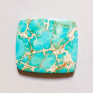 Op de afbeelding: Een rechthoekige cabochon edelsteen met afgeronde hoeken, voorzien van een levendige turquoise blauwe kleur met een complex netwerk van crème en lichtbruine matrixaders over het gepolijste oppervlak van de steen.