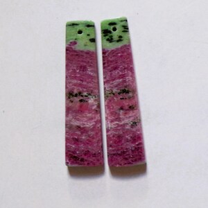 May include: Two rectangular gemstone pendants. The top portion is light green with black speckles, transitioning to a vibrant pinkish-red hue below. Each pendant has a small hole at the top for stringing. The gemstones are likely for jewelry making.