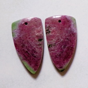 May include: Two shield-shaped gemstones with a hole drilled at the top for jewelry making. The stones have a vibrant color palette, featuring a blend of deep pink, green, and black hues. The gemstones are polished and smooth, with a natural, organic appearance.