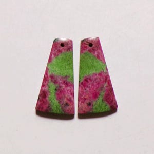 Peut inclure: Deux pendentifs en pierre de forme trapézoïdale présentant un motif marbré de couleurs magenta vif et vert lime éclatant. Chaque pièce possède une surface lisse et polie ainsi qu'un petit trou circulaire percé près du bord supérieur.