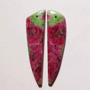 Könnte beinhalten: Zwei polierte Edelsteinanhänger. Jeder Anhänger hat eine lange, schlanke Form mit abgerundeter Oberseite und spitzer Unterseite. Die Steine weisen eine Mischung aus leuchtenden Rosa- und Grüntönen auf, mit einem Loch zum Auffädeln.