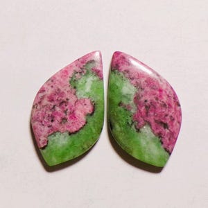 Könnte beinhalten: Zwei polierte, marquiseförmige Cabochons mit einer natürlichen Mischung aus leuchtend magentafarbenen und hellgrünen Mineralmustern. Jeder Stein zeigt eine glatte, glänzende Oberfläche mit ausgeprägten Farbvariationen und organischen Texturen.