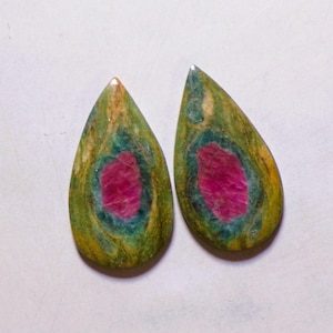 Peut inclure: Deux cabochons en forme de poire présentant un centre rose vif entouré d'anneaux concentriques bleu sarcelle profond et vert olive terreux. Ces pierres polies présentent une surface lisse et arrondie avec des variations de couleurs distinctes.
